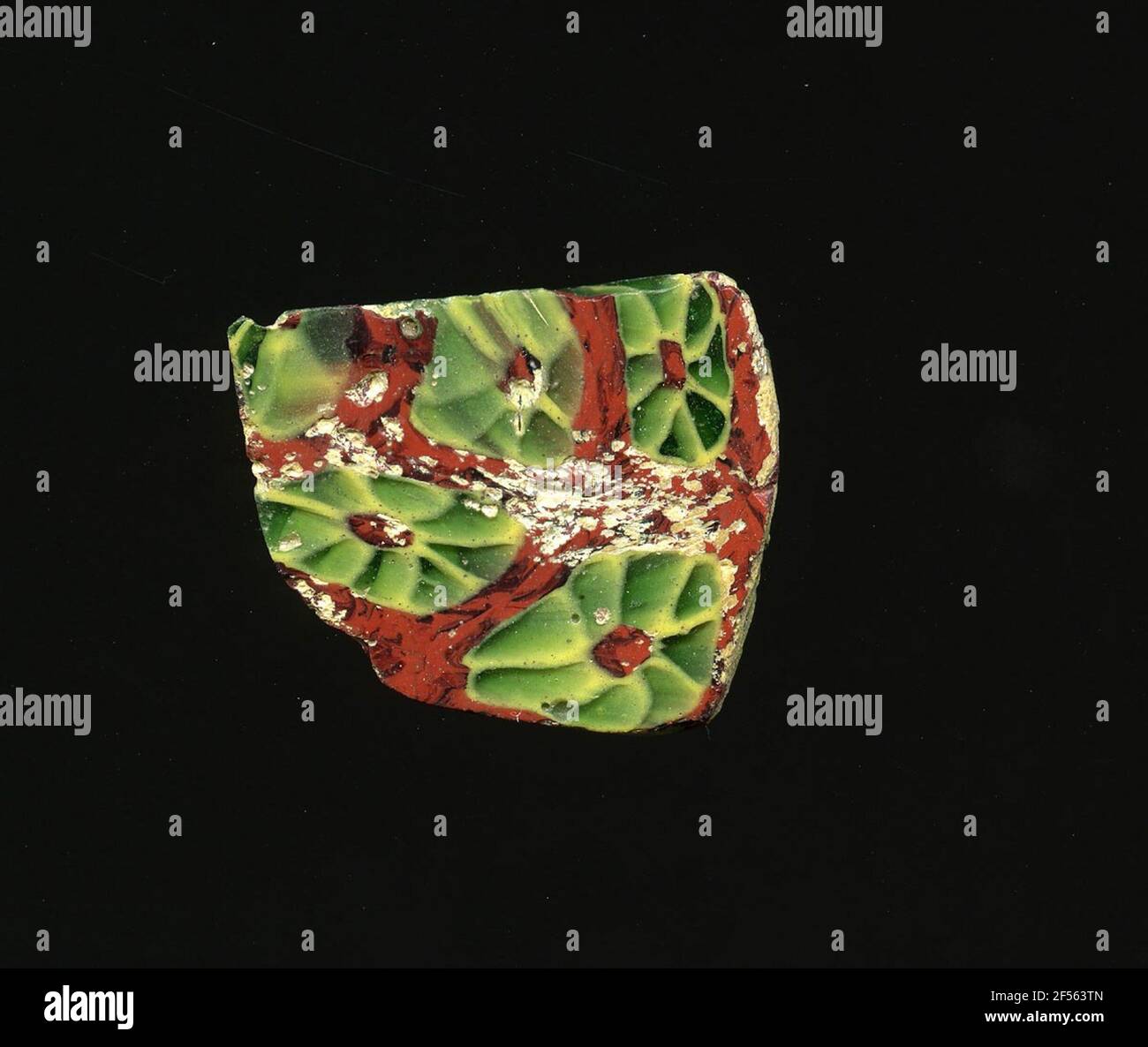 Fragment d'un vaisseau. Une faible courbure du fragment de verre permet l'affectation à un vaisseau, éventuellement à une coque. Sur la raison marbrée rouge et noire, les fleurs peuvent être vues dans la technologie millefiori. Ils se composent d'un noyau marbré rouge et noir pour poser des points verts avec un contour jaune comme des pétales. Pour la production, des verres de différentes couleurs ont été façonnés en tubes et en tiges. Elles étaient comprimées et chauffées, ce qui entraînait une longue barre. Le verre coupé en tranches de ​​now avait des motifs amorphes ou floraux différents. Par la suite, plusieurs de ces plaquettes ont été repliées ensemble, réchauffées à nouveau et pressées en forme vasculaire. Banque D'Images