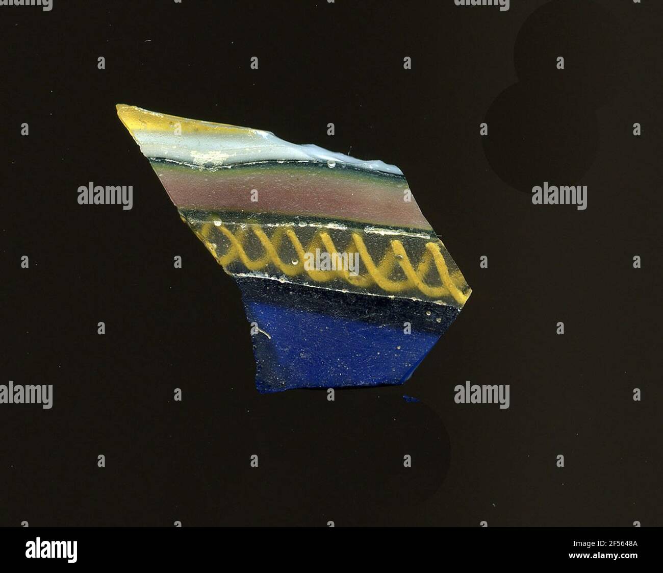 Fragment d'un vaisseau. Une faible courbure du fragment de verre permet l'affectation à un vaisseau, éventuellement à une coque. À travers le verre, plusieurs bandes de différents modèles sont dessinées en parallèle: Obtenir un bleu, un transparent avec un fil jaune en spirale, une pourriture noire avec des bords étroits verts et une bande blanche. Pour la production, des verres de différentes couleurs ont été façonnés sur des tubes et des tiges. Elles étaient comprimées et chauffées, ce qui entraînait une longue barre. Le verre coupé en tranches de ​​now avait des motifs amorphes ou floraux différents. Par la suite, plusieurs de ces plaquettes ont été pliées, chauffées à nouveau et pressées en forme vasculaire. BUR Banque D'Images