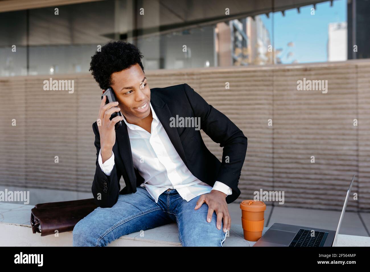 Homme souriant, homme d'affaires parlant sur un smartphone, assis sur un mur de soutènement tout en regardant loin Banque D'Images