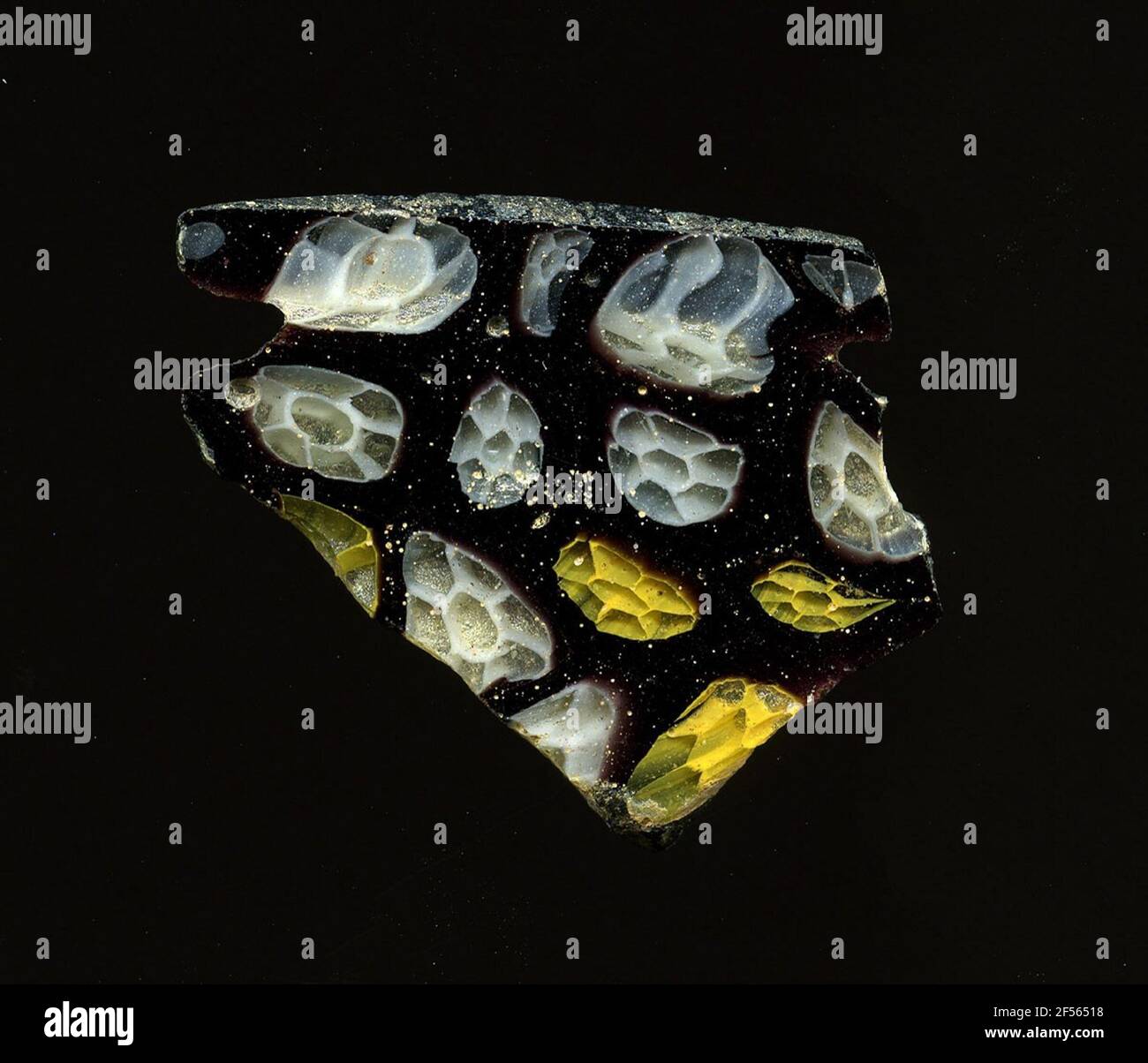 Fragment d'un vaisseau. Une faible courbure du fragment de verre permet l'affectation à un vaisseau. À cause violette, une variété de petites fleurs peuvent être vues, qui sont construites de la même façon. Les contours jaune et blanc encadrent le noyau et les feuilles de fleurs transparentes. Pour la production, des verres de différentes couleurs ont été formés en tubes et en tiges. Elles étaient comprimées et chauffées, ce qui entraînait une longue barre. Le verre coupé en tranches de ​​now avait des motifs amorphes ou floraux différents. Par la suite, plusieurs de ces plaquettes ont été fusionnées, réchauffées à nouveau et pressées en forme vasculaire. Burry pour le fabricant d'élaborer Banque D'Images