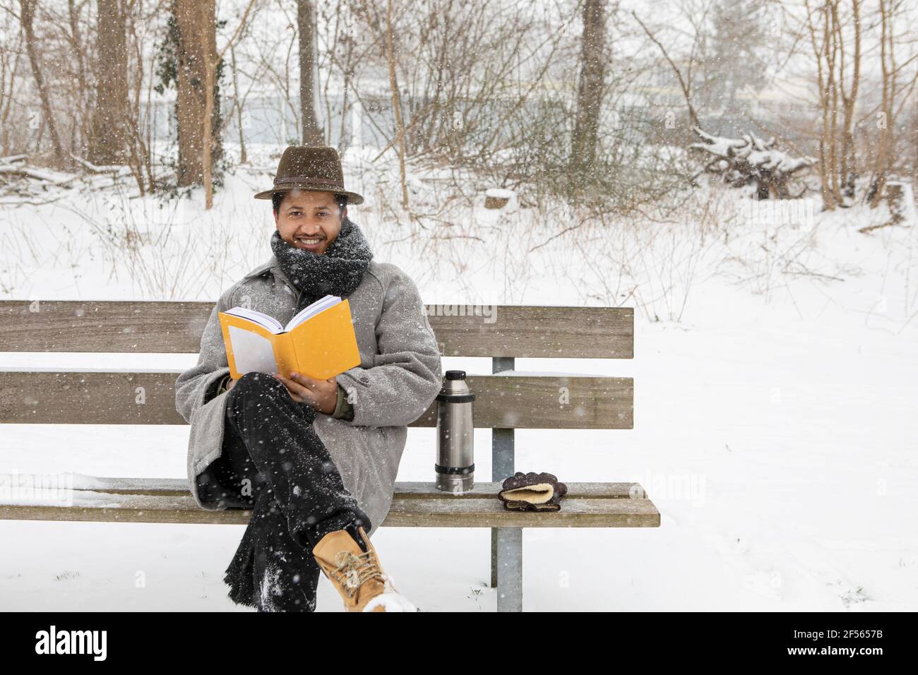 Homme souriant lisant le livre tout en étant assis sur le banc pendant l'hiver Banque D'Images