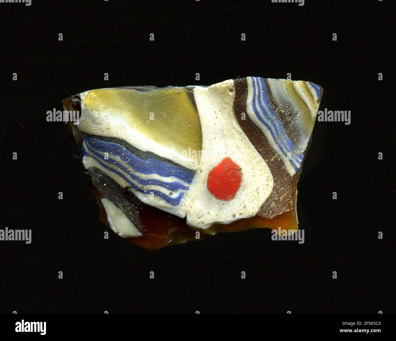 Fragment d'un vaisseau. Une faible courbure du fragment de verre permet l'affectation à un vaisseau, éventuellement à une coque. Dans le verre brun, les motifs abstraits sont reconnaissables. Le centre est une zone blanche amorphe avec un point rouge. Il y a des bandes qui tirent alternativement des bandes bleu-blanc ou vert-jaune-blanc dans différentes directions sur la bordure. Pour la production, des verres de différentes couleurs ont été formés en tubes et en tiges. Elles étaient comprimées et chauffées, ce qui entraînait une longue barre. Le verre coupé en tranches de ​​now avait des motifs amorphes ou floraux différents. Par la suite, plusieurs de ces plaquettes ont été pliées ensemble, lui Banque D'Images