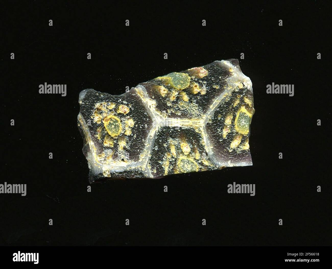 Fragment d'un vaisseau. Une faible courbure du fragment de verre permet l'affectation à un vaisseau. Le verre rouge foncé traverse une grille blanche qui rappelle les nids d'abeilles. Dans les champs, on peut voir une floraison dans la technologie millefiori. Sur un point vert avec une bordure jaune, les petits points jaunes se posent sous forme de pétales. Pour la production, des verres de différentes couleurs ont été moulés dans des tubes et des tiges. Elles étaient comprimées et chauffées, ce qui entraînait une longue barre. Le verre coupé en tranches de ​​now avait des motifs amorphes ou floraux différents. Par la suite, plusieurs de ces plaquettes ont été fusionnées, réchauffées à nouveau et formées. Burry pour l'e Banque D'Images