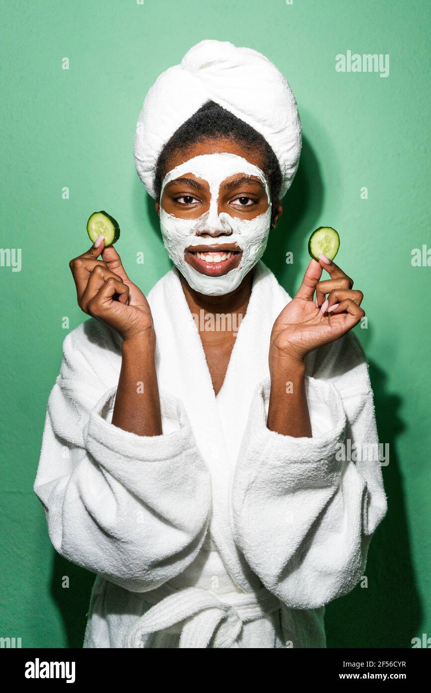 Jeune femme avec masque facial tenant le concombre tout en se tenant contre arrière-plan vert Banque D'Images