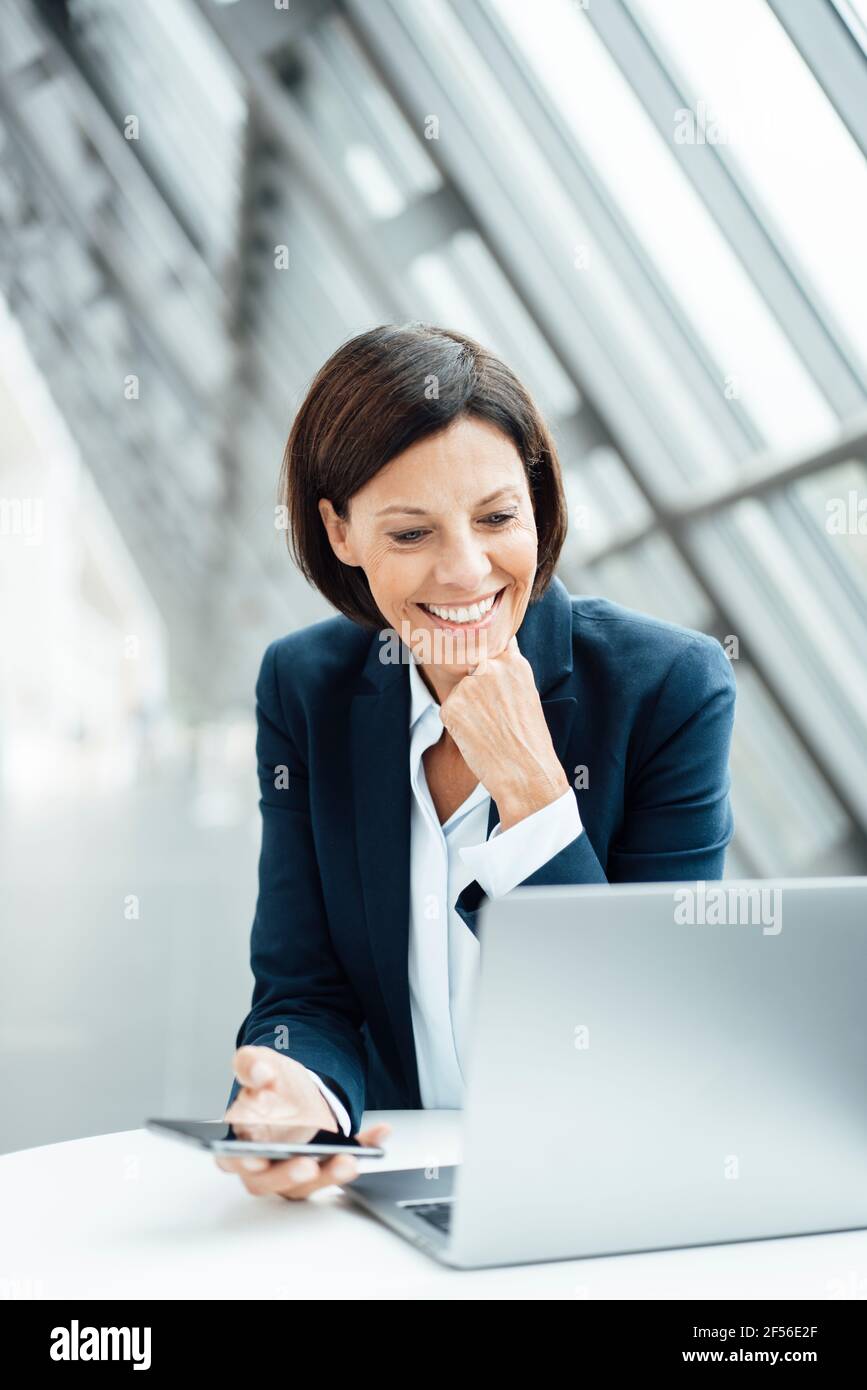 Femme d'affaires souriante, main sur le menton, travaillant sur un ordinateur portable couloir Banque D'Images
