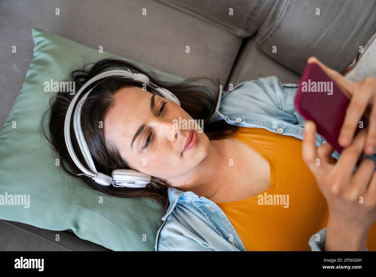 Jeune femme avec un casque utilisant un téléphone portable pendant qu'elle est allongée canapé à la maison Banque D'Images