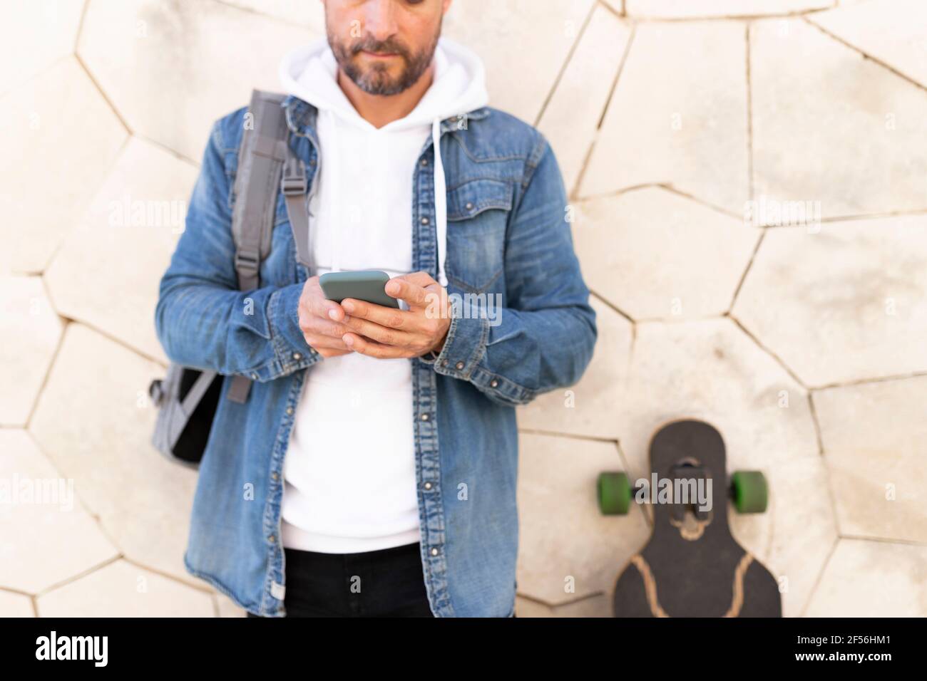 Homme avec un skateboard utilisant un smartphone tout en se penchant sur le mur Banque D'Images