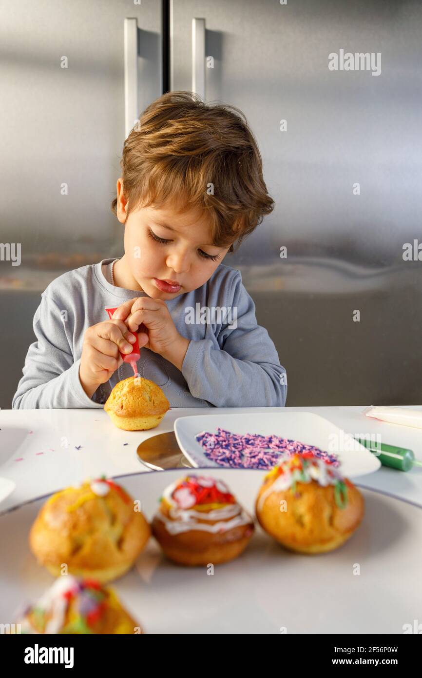 Une adorable petite fille léchant le doigt tout en décorant un muffin au comptoir de la cuisine Banque D'Images