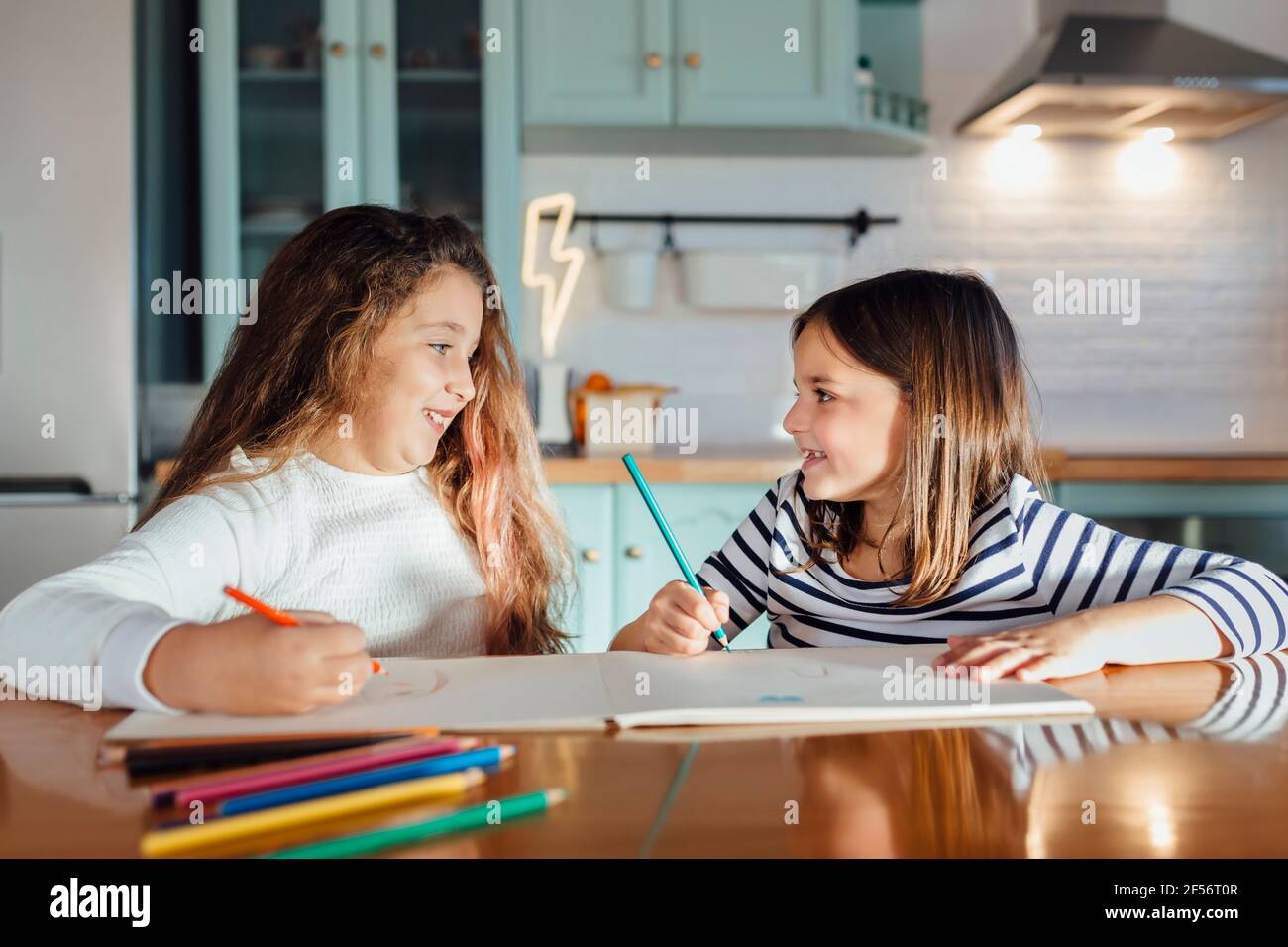 Souriantes filles regardant les unes les autres tout en colorant avec de la couleur crayon sur papier tout en étant assis à la table dans la cuisine Banque D'Images