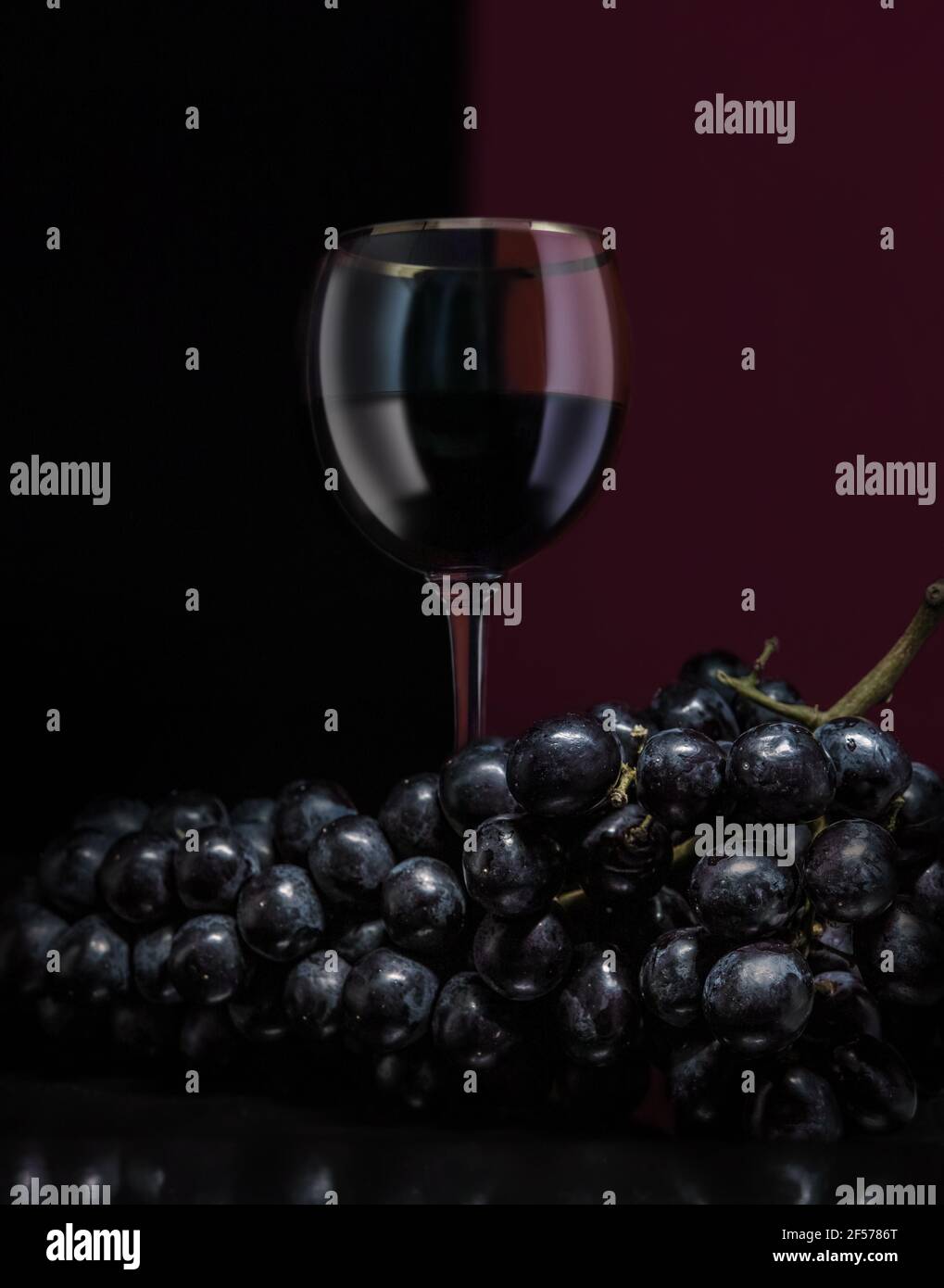 Un verre de vin rouge derrière un bouquet de raisins noirs. L'arrière-plan est à moitié rouge très foncé, à moitié noir. Banque D'Images