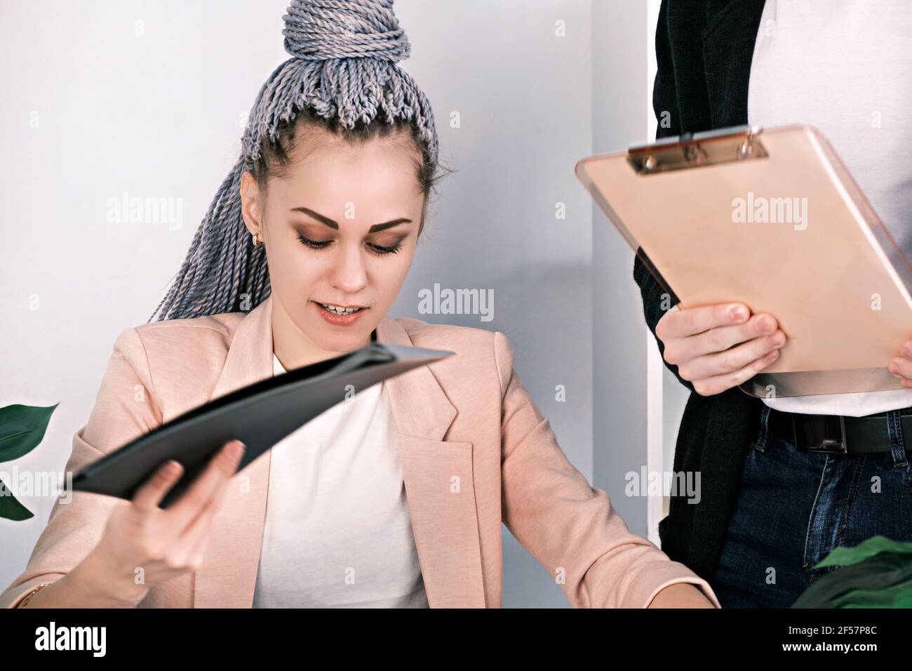Jeune femme moderne avec des dreadlocks de canecalon ou des queues de cochon tient des dossiers de documents assis dans le bureau. L'homme se tient à côté d'elle avec un presse-papiers dans Banque D'Images