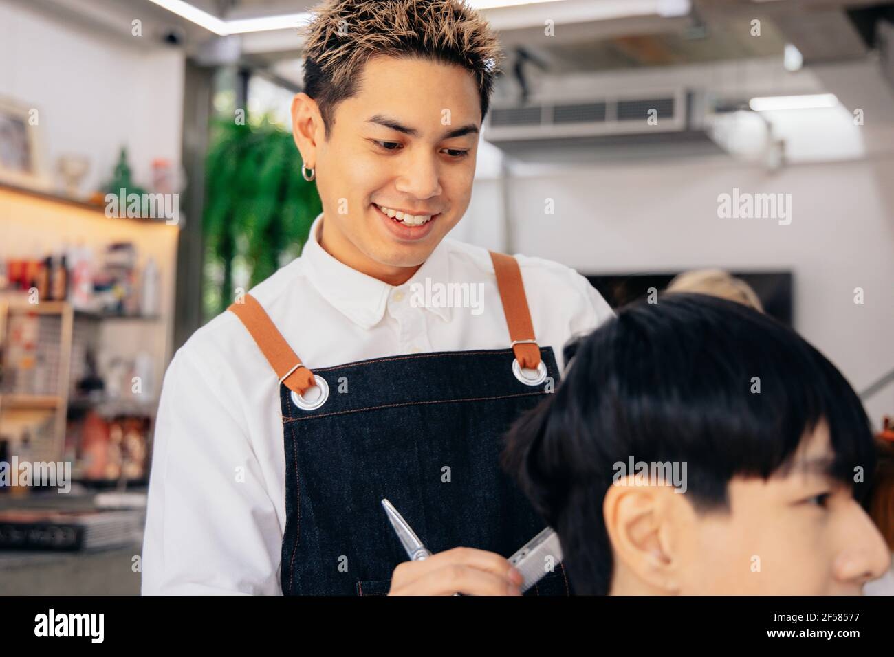 Joyeux coiffeur de jeune asiatique souriant portant un tablier dans moderne salon tenant la brosse et les ciseaux de couper les cheveux de client Banque D'Images