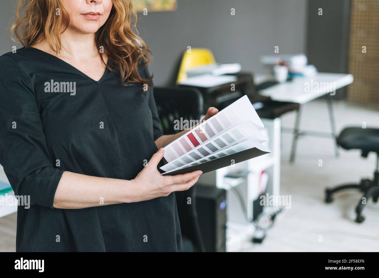 Charmante femme adulte d'âge moyen avec un architecte de cheveux bouclés avec palette de couleurs à portée de main dans un environnement de travail moderne Banque D'Images
