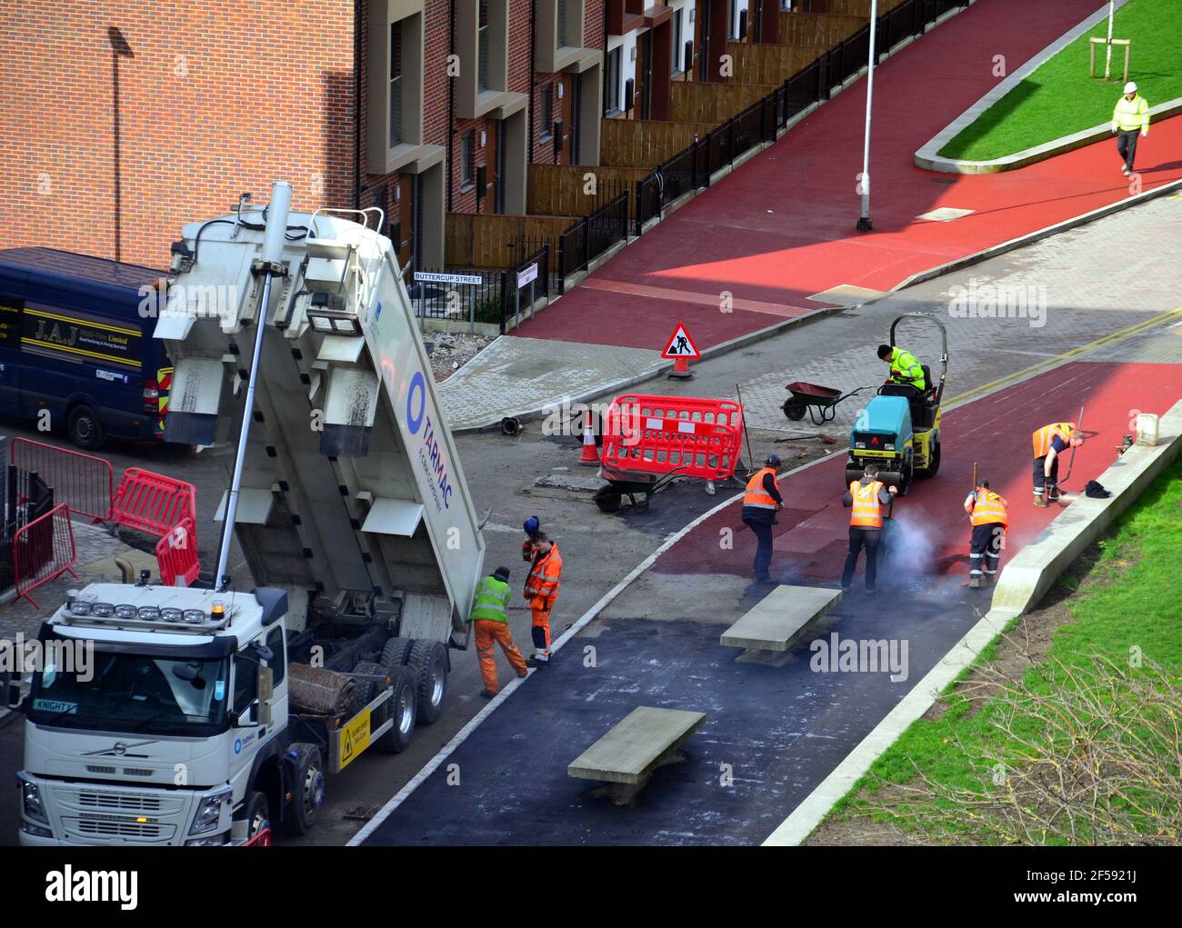 Vue aérienne des travailleurs qui ponent du tarmac rouge ou de l'asphalte rouge ou du bitume rouge ou du goudron rouge pour créer un pavé sur une nouvelle route dans la région de Brunswick à Ardwick, Manchester, Angleterre, Royaume-Uni. Les ouvriers utilisent des râteaux pour mettre le tarmac à niveau, car un petit rouleau attend de l'écraser sur une surface lisse. Un camion à côté fournit plus d'asphalte rouge. Banque D'Images