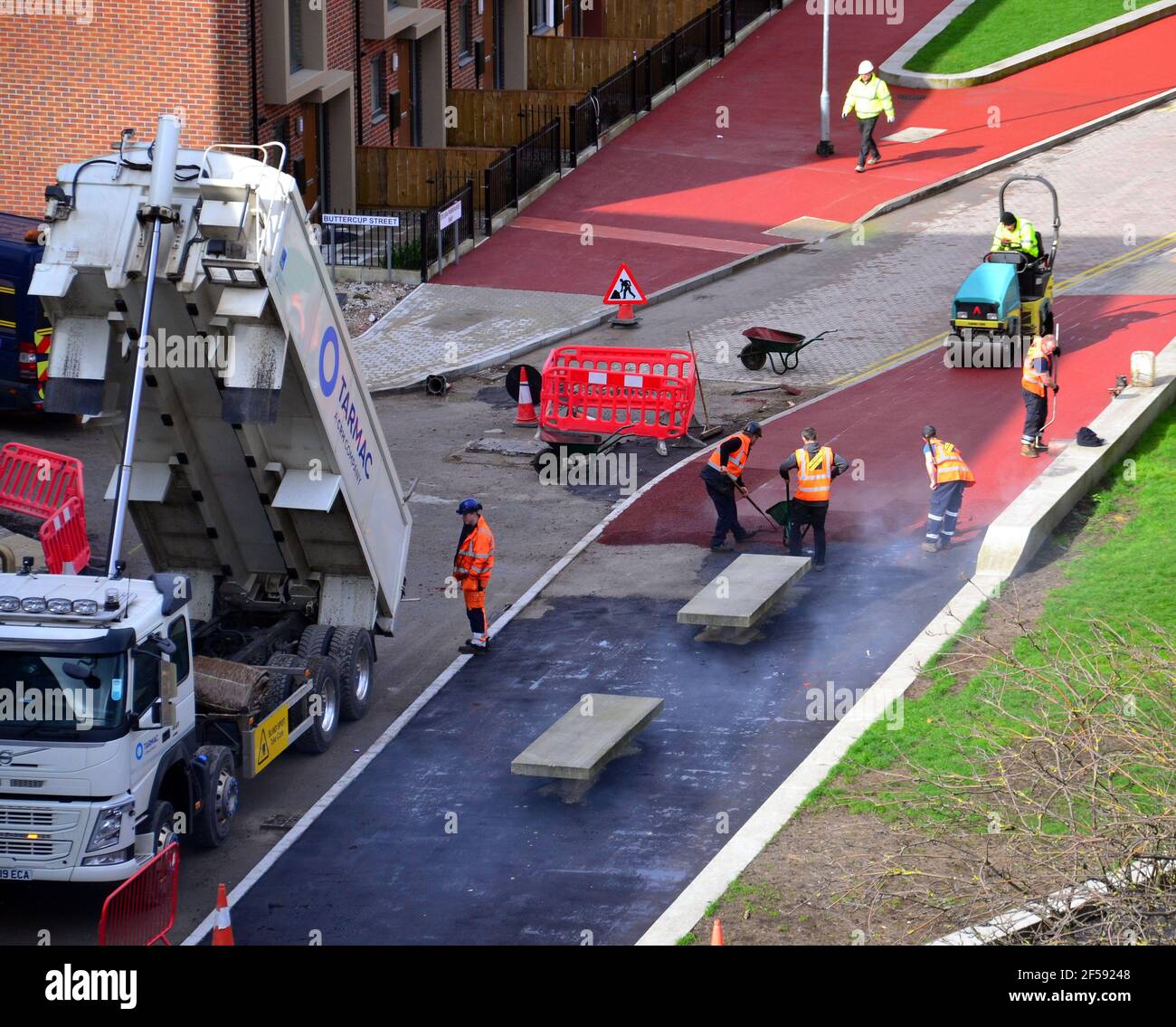 Vue aérienne des travailleurs qui ponent du tarmac rouge ou de l'asphalte rouge ou du bitume rouge ou du goudron rouge pour créer un pavé sur une nouvelle route dans la région de Brunswick à Ardwick, Manchester, Angleterre, Royaume-Uni. Les ouvriers utilisent des râteaux pour mettre le tarmac à niveau, car un petit rouleau attend de l'écraser sur une surface lisse. Un camion à côté fournit plus d'asphalte rouge. Banque D'Images