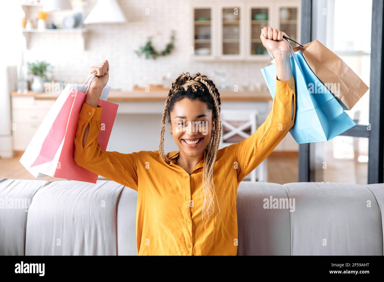 Bonne jeune femme afro-américaine branchée, heureuse de ses nouveaux achats, assise sur le canapé à la maison, tenant des sacs en papier avec de nouveaux vêtements dans ses mains, regardant l'appareil photo, souriant heureux Banque D'Images