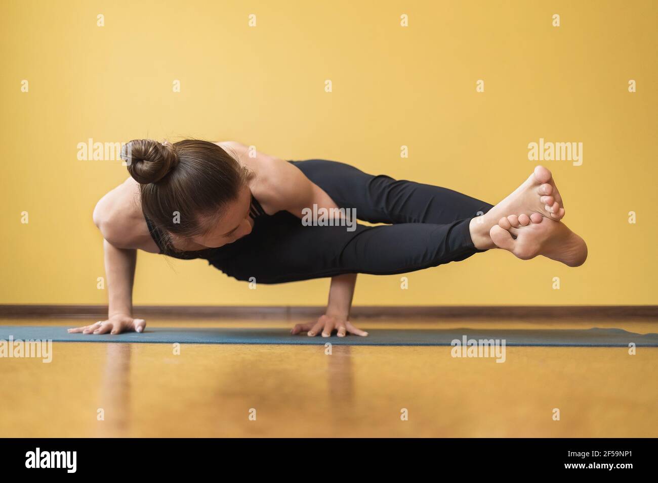 Une femme dans un survêtement noir d'une seule pièce exécute l'exercice Ashtavakrasana, une main contre le fond du mur. Banque D'Images