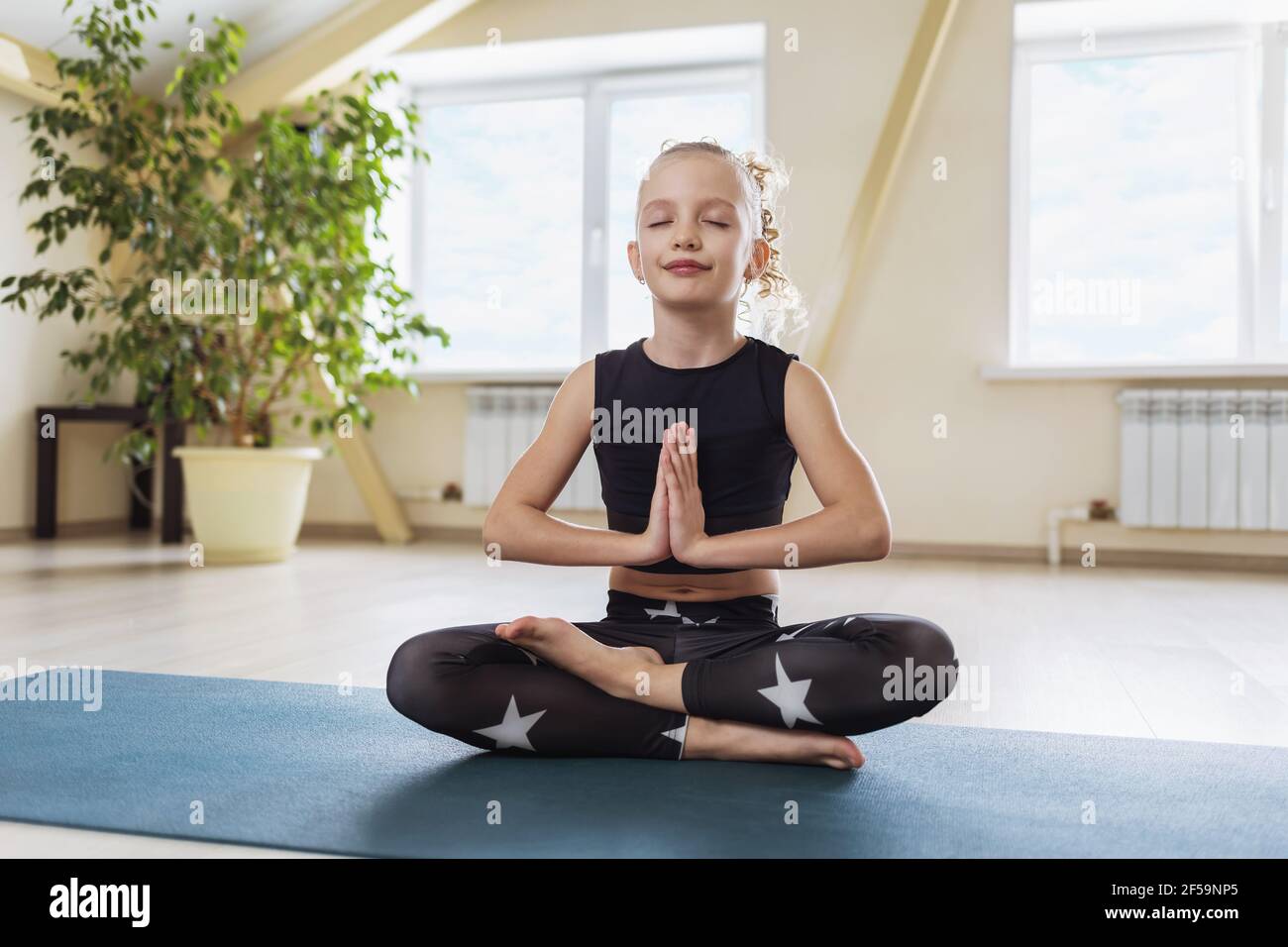 Une petite fille, dans des vêtements de sport noirs, pratiquant le yoga, est engagée dans la méditation, assise dans la position lotus, montrant avec ses mains le salut de n Banque D'Images