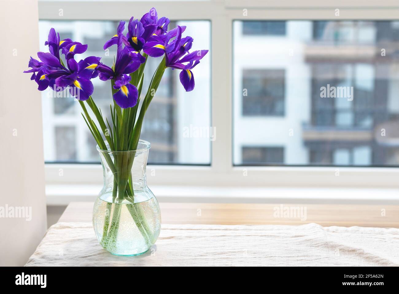 Bouquet d'iris violets dans un vase en verre transparent une nappe de linge sur une table en bois près de la fenêtre dans une cuisine moderne et lumineuse Banque D'Images