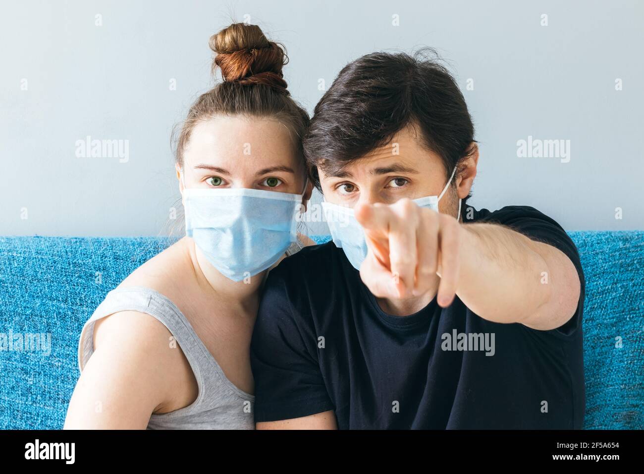 Couple dans les masques médicaux bleus assis sur le canapé, regardant l'appareil photo et il pointant le doigt vers l'appareil photo Banque D'Images