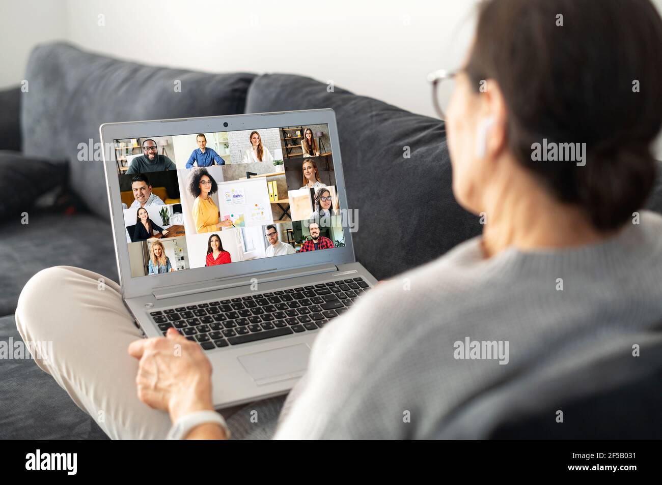 Femme contemporaine sénichée sur le canapé confortable et utilisant un ordinateur portable pour la connexion vidéo avec un groupe de personnes diverses, vue arrière d'une femme plus âgée impliquée webinar, vidéo conférence Banque D'Images