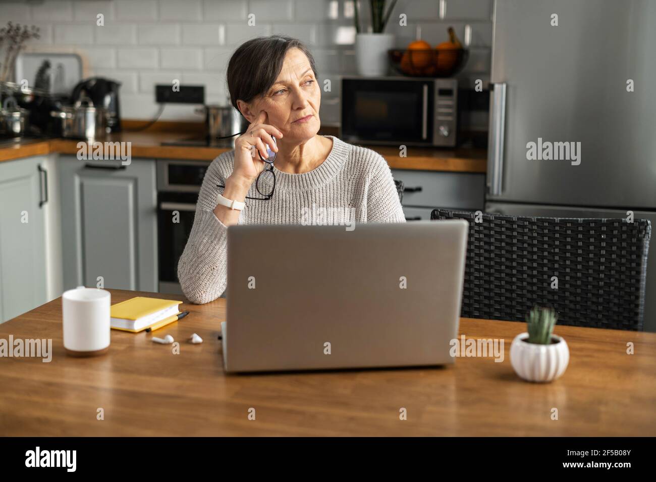 Femme pensive à la retraite utilisant un ordinateur portable pour le travail à distance de la maison, la femme senior s'assoit avec un ordinateur au bureau de cuisine, regarde loin et pense Banque D'Images