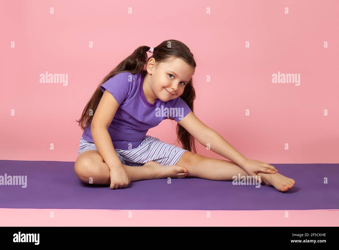 portrait complet d'une fille souriante effectuant des exercices matinaux et des coudes latéraux pour plus de flexibilité assis sur un tapis de sport violet, isolé sur fond rose Banque D'Images