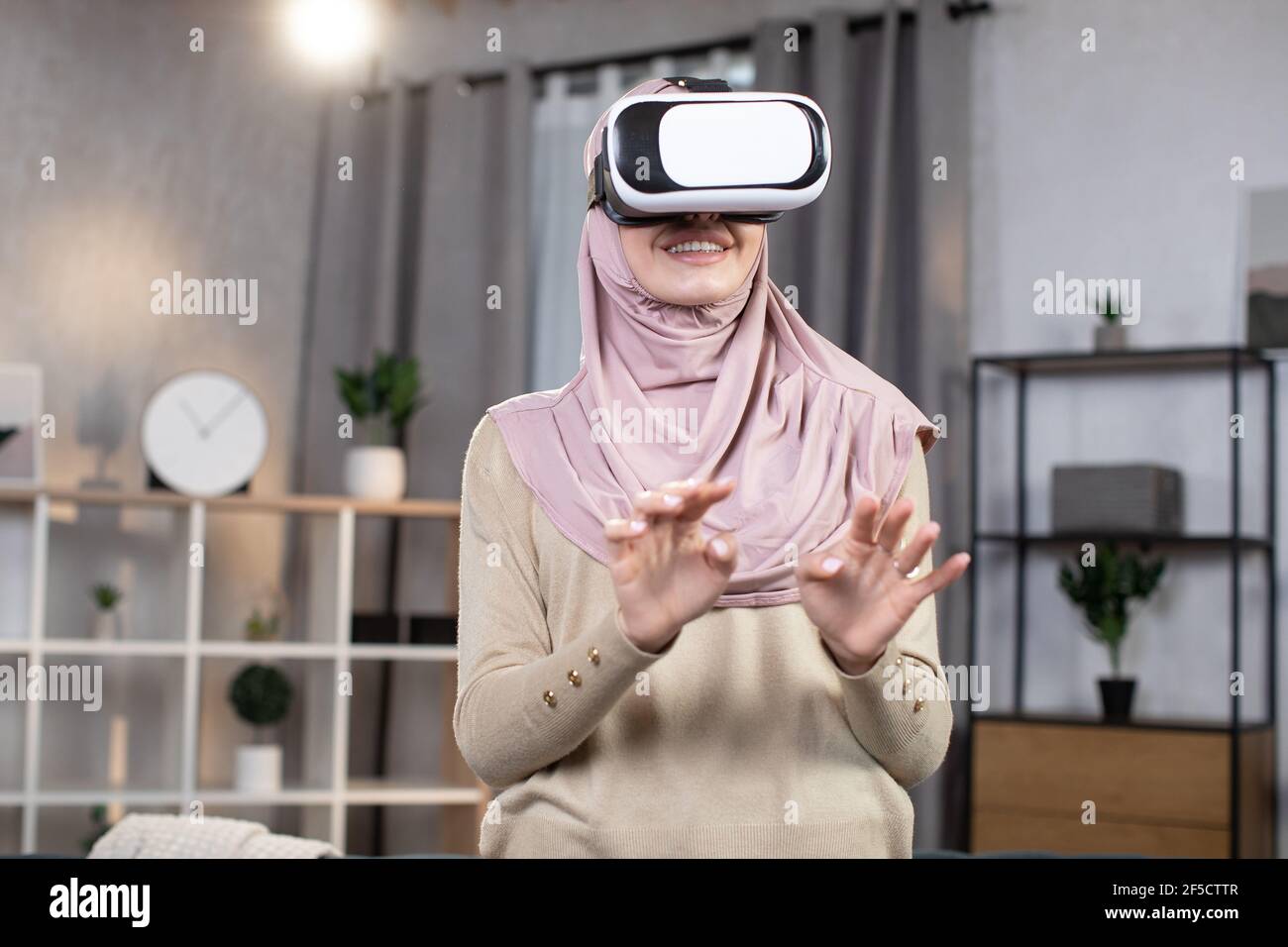 Jeune femme musulmane dans le hijab, s'amuser dans le casque de réalité  virtuelle, jouer à un jeu vidéo interactif, gesturant dans les airs, tout  en se tenant dans le salon confortable moderne