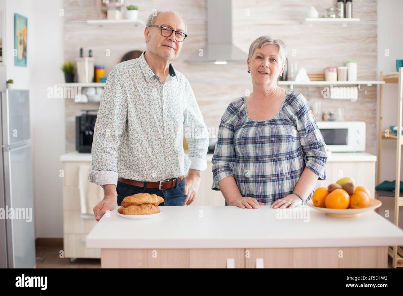 Portrait d'un vieux couple gai regardant dans l'appareil photo dans la cuisine tout en préparant le petit déjeuner. Bonne femme et homme à la retraite. Banque D'Images