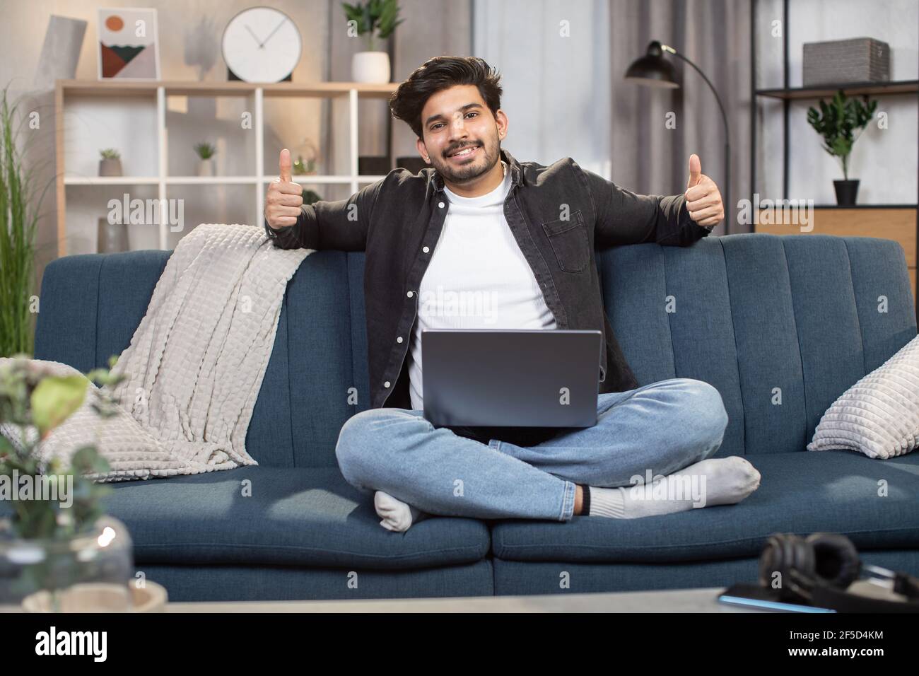 Charmant Indien de 30 ans souriant, assis sur un canapé confortable à la maison, se sentant détendu tout en passant son temps libre avec un ordinateur portable, montrant ses pouces à l'appareil photo. Banque D'Images
