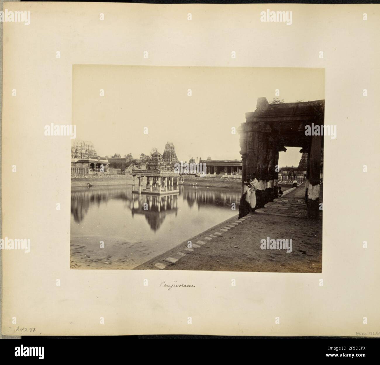 Conjivoram. Vue sur le réservoir d'un temple de Kanchipuram. Au centre de la piscine se trouve un kiosque avec des piliers sculptés et un dôme en forme de cloche sculpté ornement sur le toit. Un homme se tient sur le côté droit de l'image enveloppé dans un tissu avec un livre dans ses mains, et un deuxième homme s'assoit sur la marche supérieure du stepwell qui entoure la piscine. Les bâtiments, les piliers, les porches en pierre et les gopuras sont visibles le long des côtés et en arrière-plan. (Recto, montage) en bas à gauche, manuscrit au crayon: 'A42.82 (Bou)' en bas au centre, manuscrit à l'encre noire: 'Conjivoram' Banque D'Images