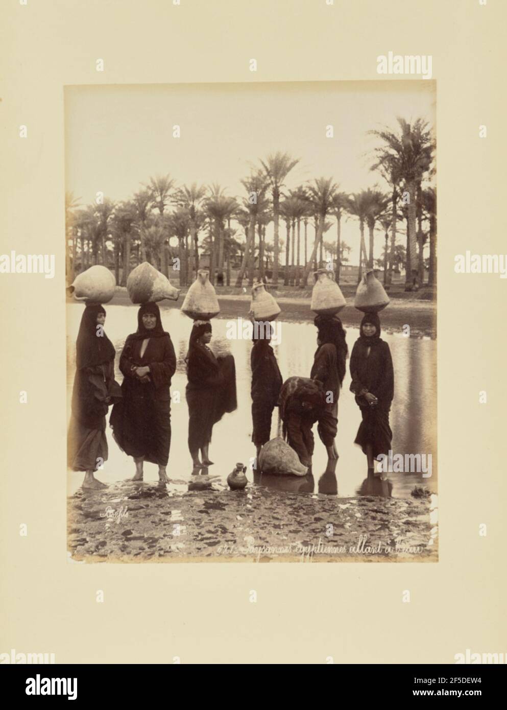 Paysannes égyptiennes sic altant à l'eau. Portrait de groupe de sept femmes autochtones debout dans l'eau, dont la plupart portent également des bateaux d'eau sur leur tête. En arrière-plan se trouvent une bosquet de palmiers et un certain nombre de bâtiments. (Recto, print) en bas à droite, inscrit en négatif: '621 _ Paysannes Ègyptiennes sic alant ‡ l'eau'; Banque D'Images