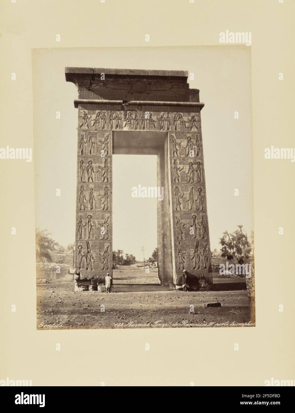 Karnak. Temple de Ramessès IV Porte triomphale. Vue sur une porte monumentale sculptée de hiéroglyphes. Le coin supérieur droit de la porte semble être manquant. Deux hommes autochtones se tiennent près de l'ouverture de la porte. (Recto, print) en bas à droite, inscrit dans le négatif: '133. Karnak. Temple de RamessËs IV. porte sic triomphale partiellement rognée'; Banque D'Images