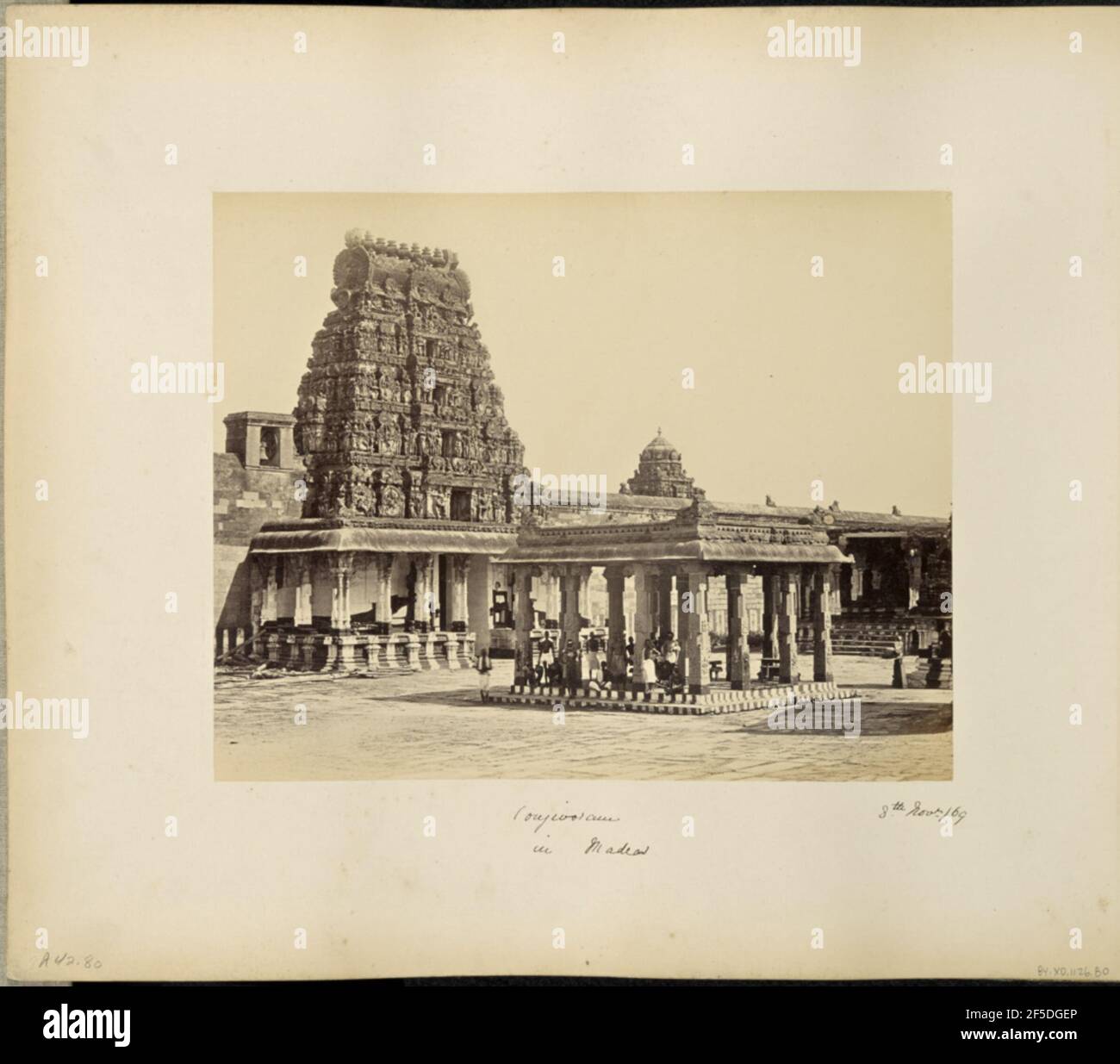 Conjivoram à Madras. Vue sur un kiosque en colonnes dans la cour d'un complexe de temples de Kanchipuram. Une gopura, ou pyramide de la porte du temple, se trouve à gauche du kiosque entouré d'un porche en pierre soutenu par des piliers sculptés. La gopura présente cinq niveaux couverts de statues sculptées ornalement de divinités hindoues. Un groupe d'hommes se rassemble sous le toit du kiosque, assis et debout, et face à la caméra. Des bandes peintes couvrent la base du kiosque et le porche du temple. (Recto, montage) en bas à gauche, manuscrit au crayon: 'A42.80 (Bou)' en bas au centre, manuscrit à l'encre noire: 'Conjivoram / in Madras' en bas r Banque D'Images