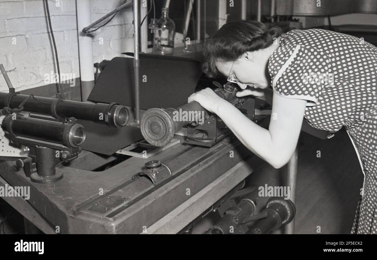 années 1950, historique, une femme porte une robe à pois ou pois dans une usine d'ingénierie légère regardant à travers une petite fenêtre sur une tige métallique en fonte, probablement en vérifiant une lentille de verre. Banque D'Images