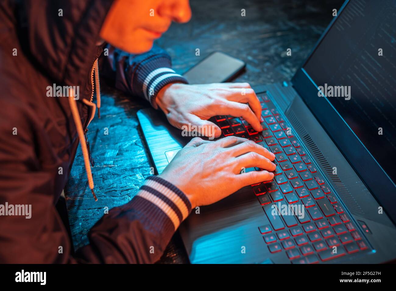 Un hacker à capuchon tape sur un clavier d'ordinateur portable dans une pièce sombre sous une lumière de néon. Le concept de la cybercriminalité et du vol d'identité Banque D'Images