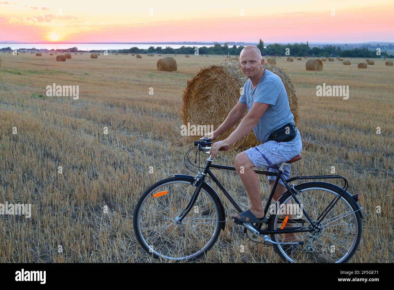 Un homme adulte sur une bicyclette sur un terrain faubouré à côté d'un secoueur au coucher du soleil. Sports et activités de vacances à la campagne. Banque D'Images