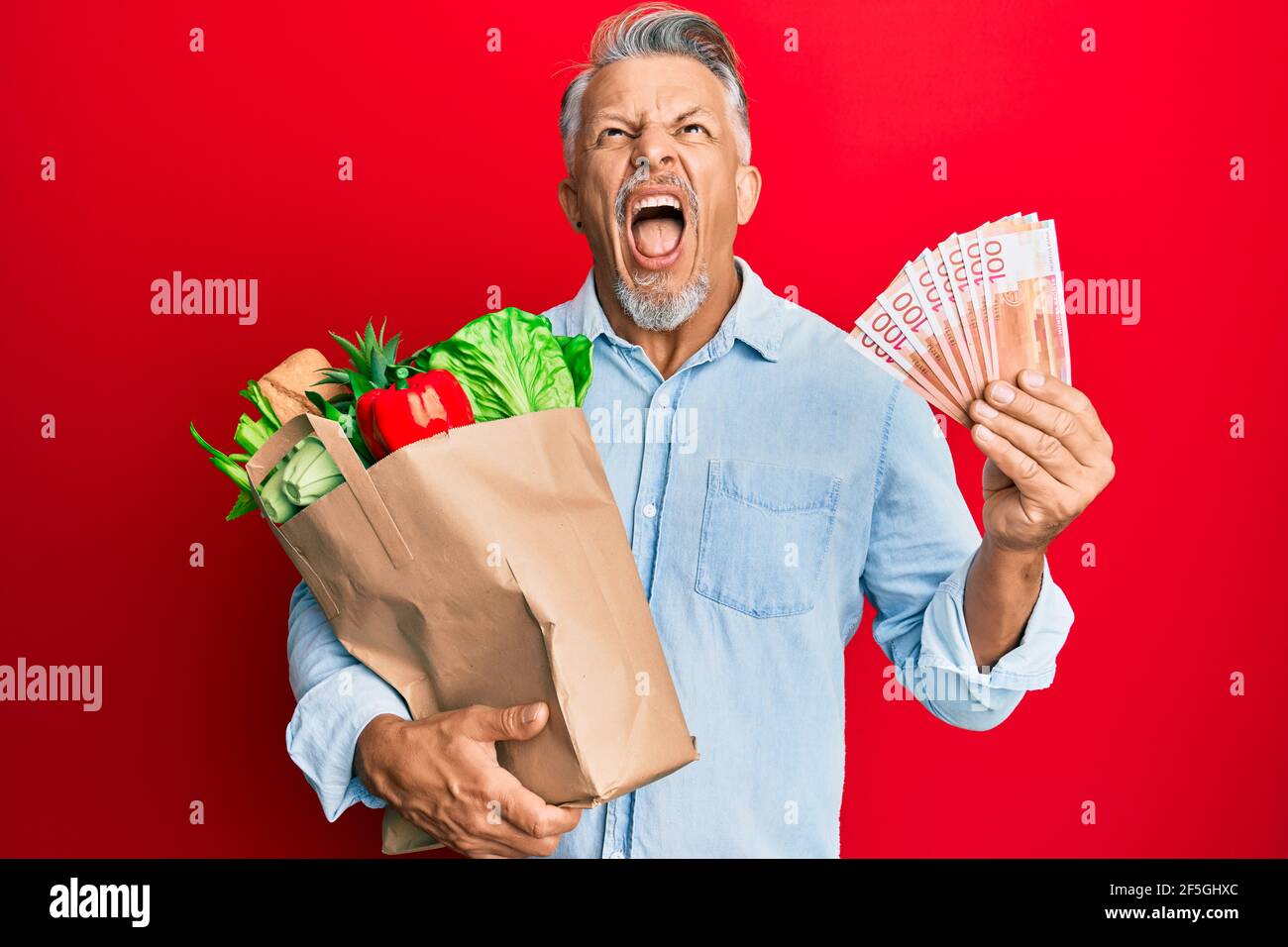 Homme à poil gris d'âge moyen tenant des provisions et des billets de banque en dollars néo-zélandais en colère et fous hurlant frustré et furieux, criant avec des toilettes de colère Banque D'Images