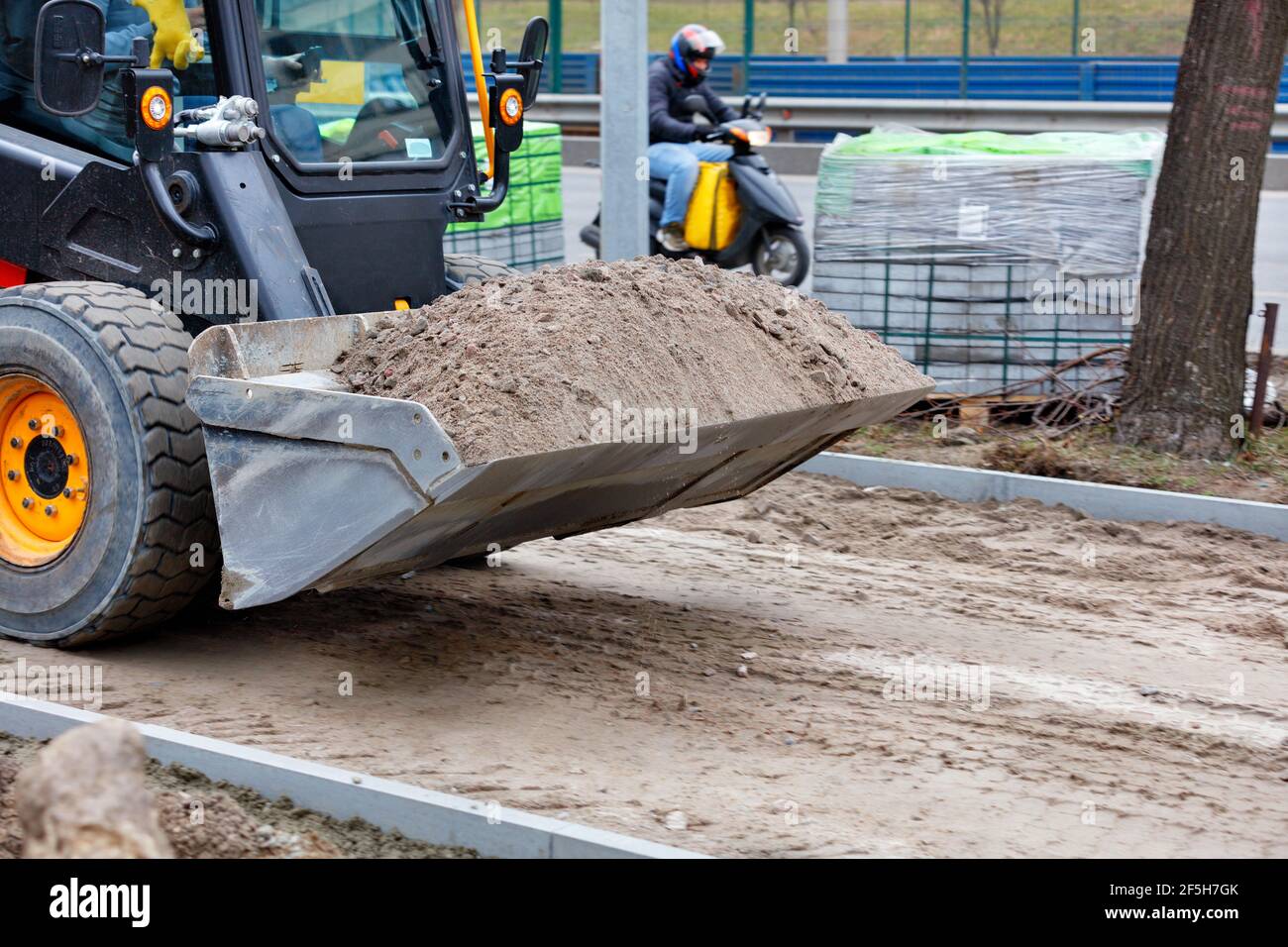 Une petite niveleuse se déplace le long d'un trottoir en construction et porte un seau de sable sur la toile de fond d'une route de la ville. Banque D'Images