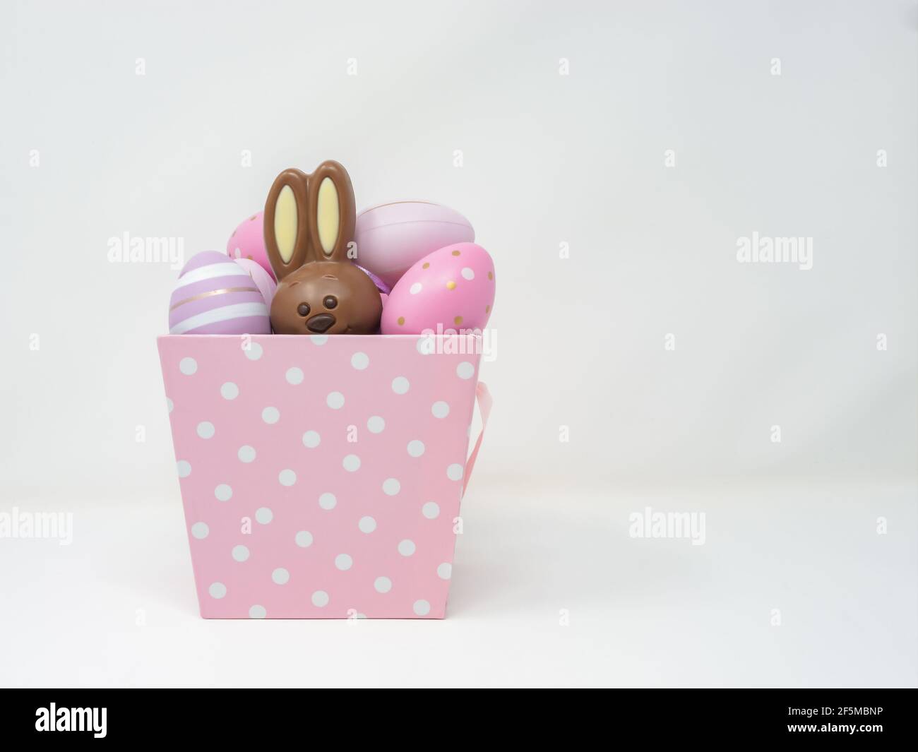 Panier rose à pois blancs rempli d'œufs décoratifs avec un lapin de Pâques en chocolat caché à l'intérieur. Arrière-plan horizontal blanc Banque D'Images