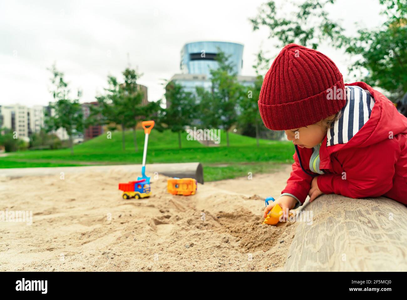 Petit enfant moderne jouant dans le bac à sable avec des jouets à l'extérieur sur le fond de la vue de paysage urbain. Banque D'Images