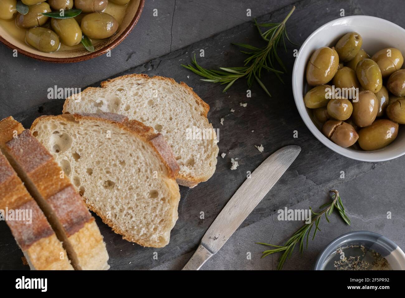 Pain de ciabatta en tranches italiennes sur planche à découper avec herbes, huile extra vierge et olives marinées sur fond de grunge foncé Banque D'Images
