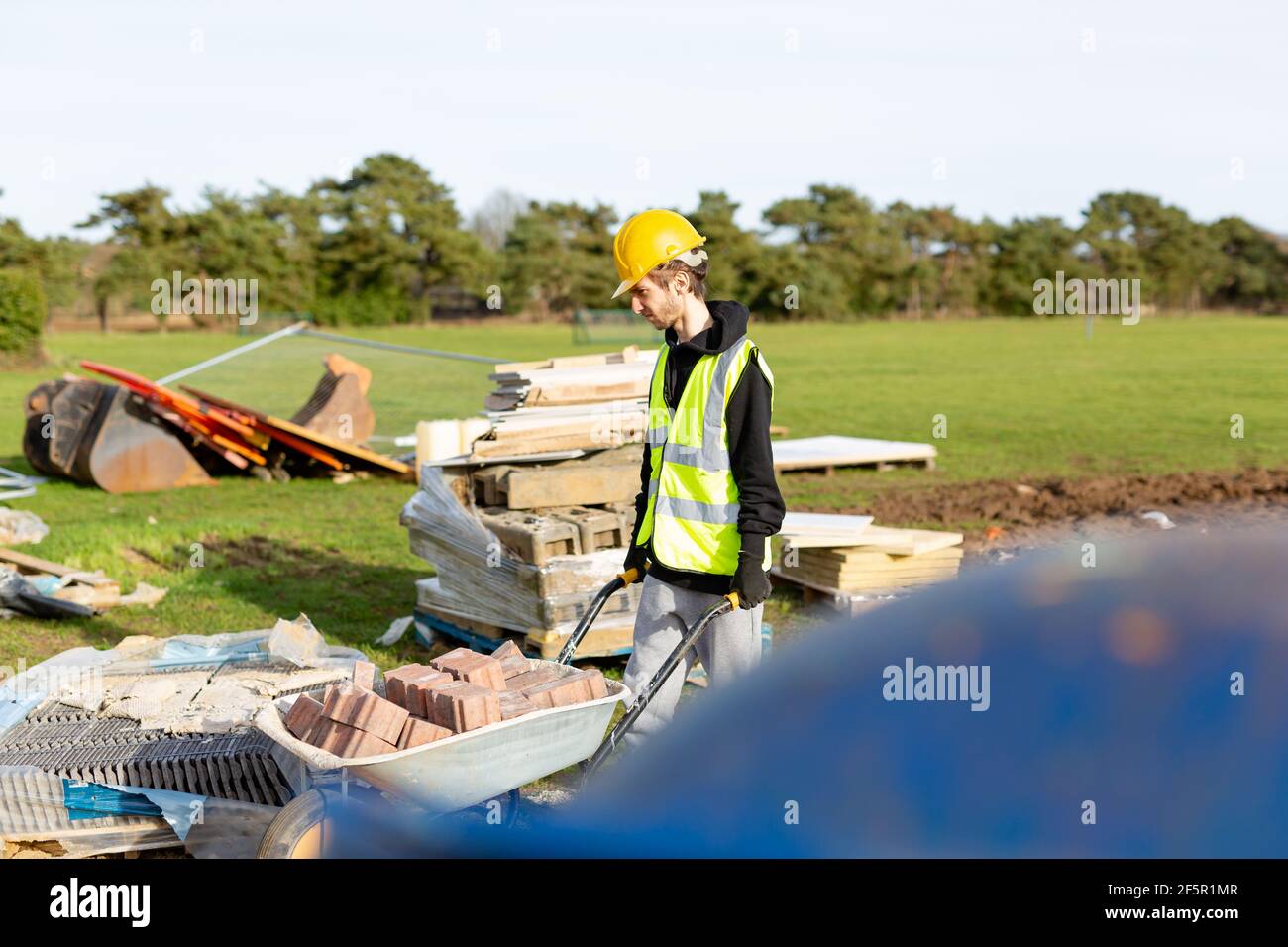 Un jeune homme de construction adulte portant un gilet haute visibilité et casque dur poussant une brouette pleine de briques pendant sur un chantier Banque D'Images