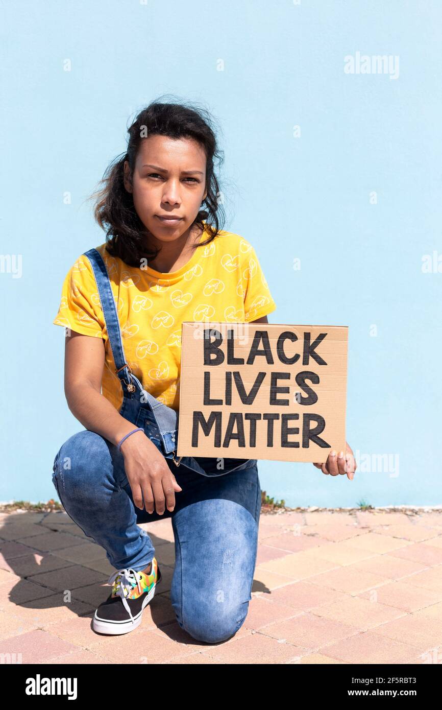 Portrait d'une femme latino-américaine avec une attitude sérieuse tenant une bannière avec le slogan Black Lives Matter. Elle s'agenouille à côté d'un mur bleu clair. Spa Banque D'Images