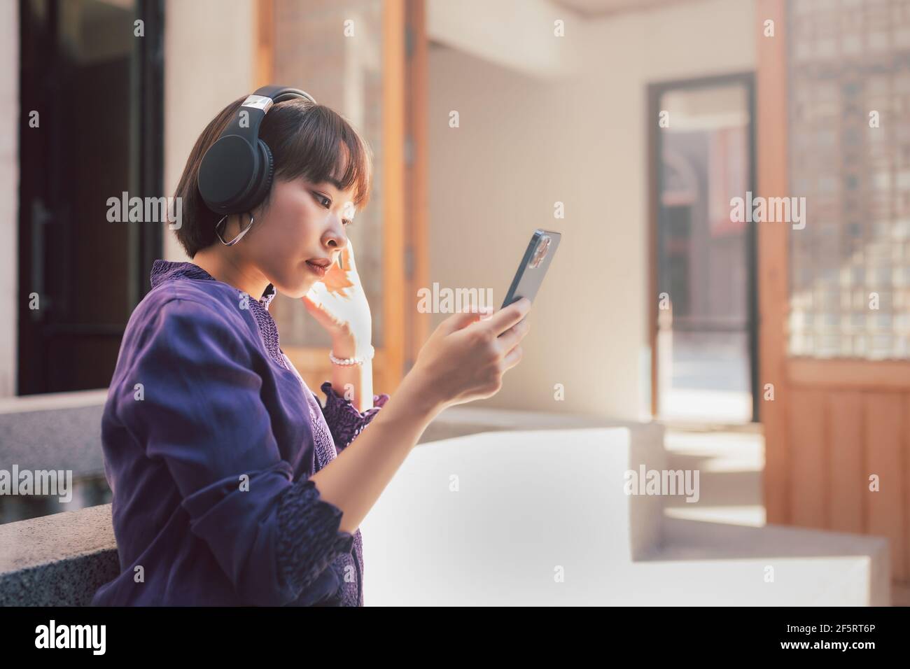 Une jeune femme asiatique heureuse écoute de la musique avec un casque via un smartphone et s'amuse en étant assise dans la rue. Banque D'Images Une jeune femme asiatique heureuse écoute de la musique avec un casque via un smartphone et s'amuse en étant assise dans la rue. Banque D'Images