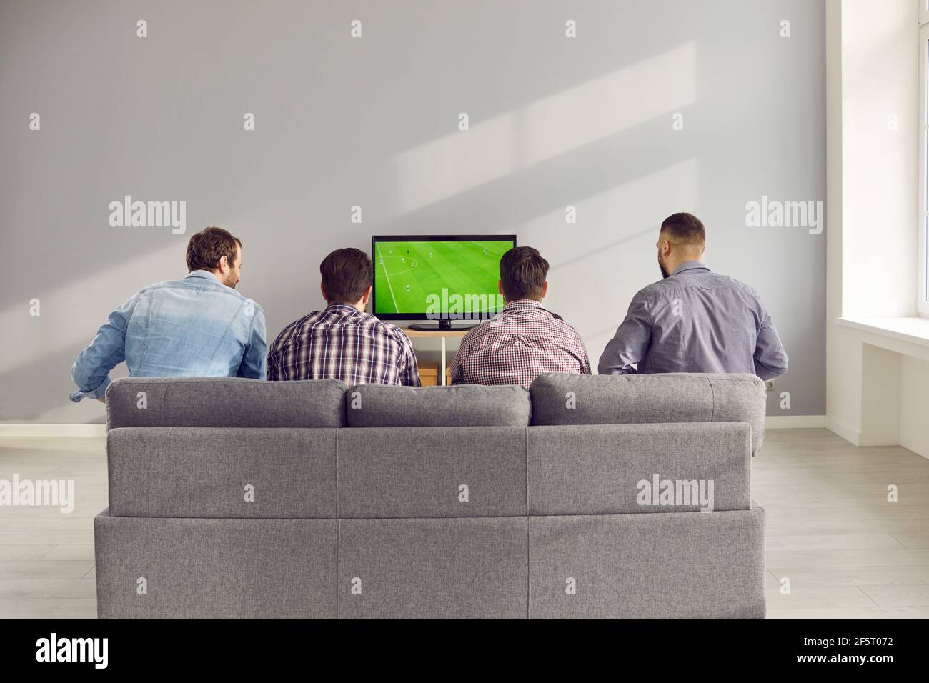 Quatre amis adultes de sexe masculin regardent un match de football à la télévision à la maison, assis sur le canapé. Banque D'Images