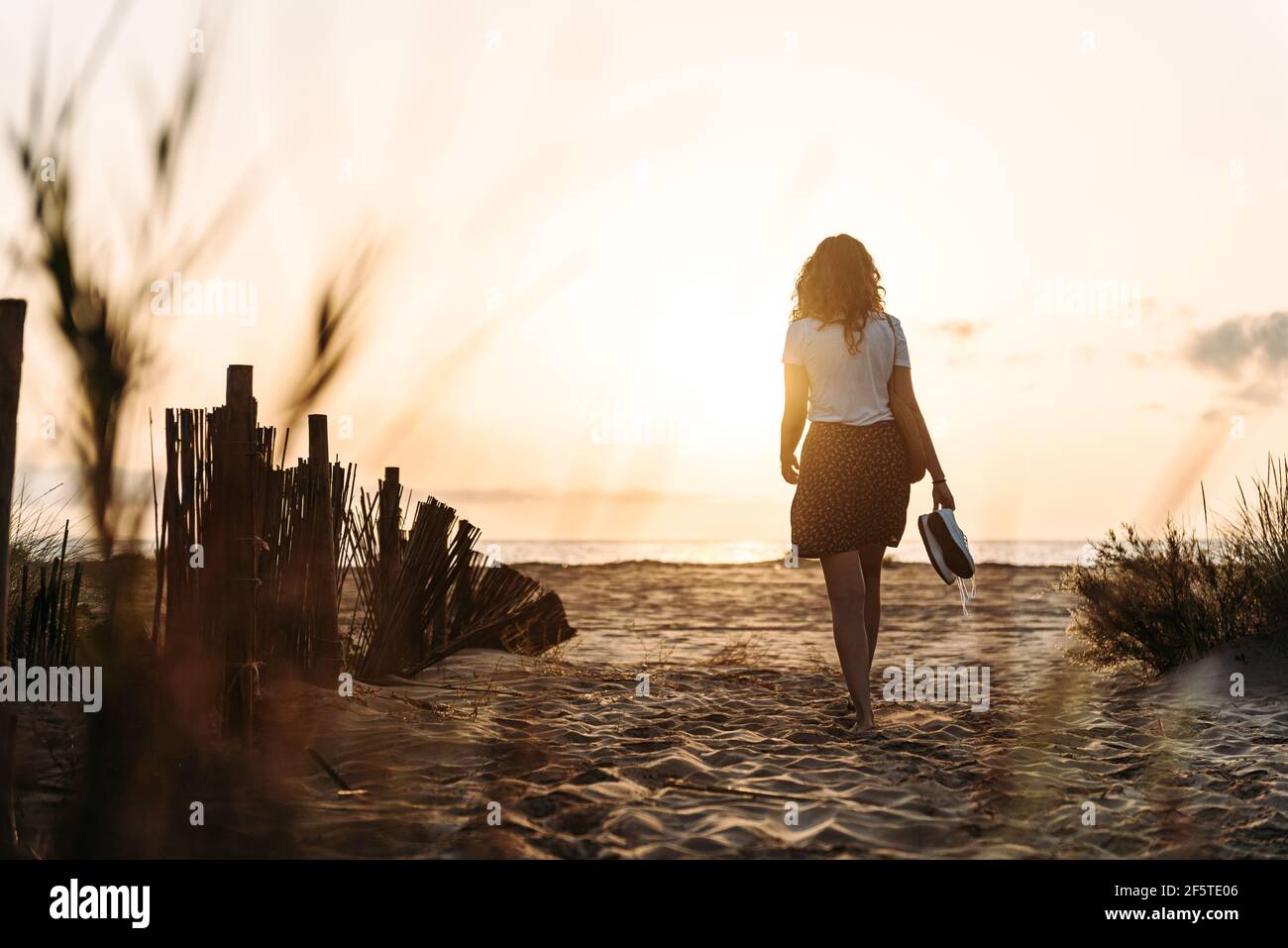 Vue arrière d'une femme méconnue en vêtements d'été qui se dirige vers mer le long de la plage de sable sur fond de ciel ensoleillé Banque D'Images