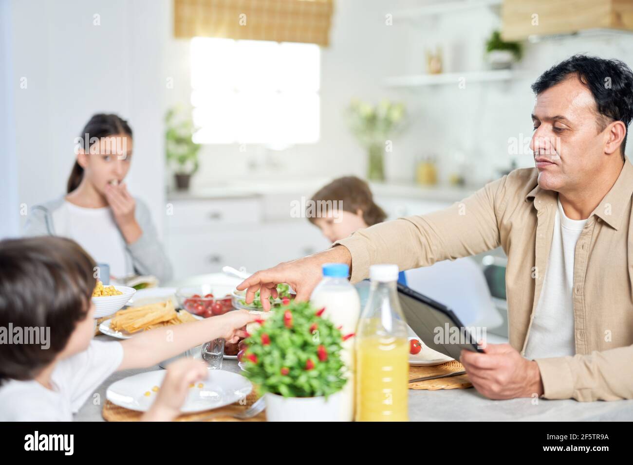 Père d'âge moyen qui apprécie un repas avec sa famille à l'aide d'une tablette numérique tout en étant assis à la table dans la cuisine à la maison Banque D'Images