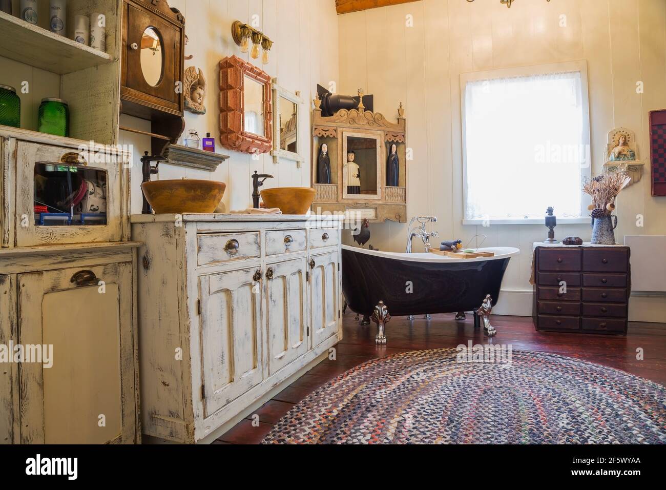Armoire de magasin générale en bois antique blanchi avec deux bols moulés lavabos et baignoire sur pieds dans la salle de bains principale avec douche tapis ovale multicolore Banque D'Images