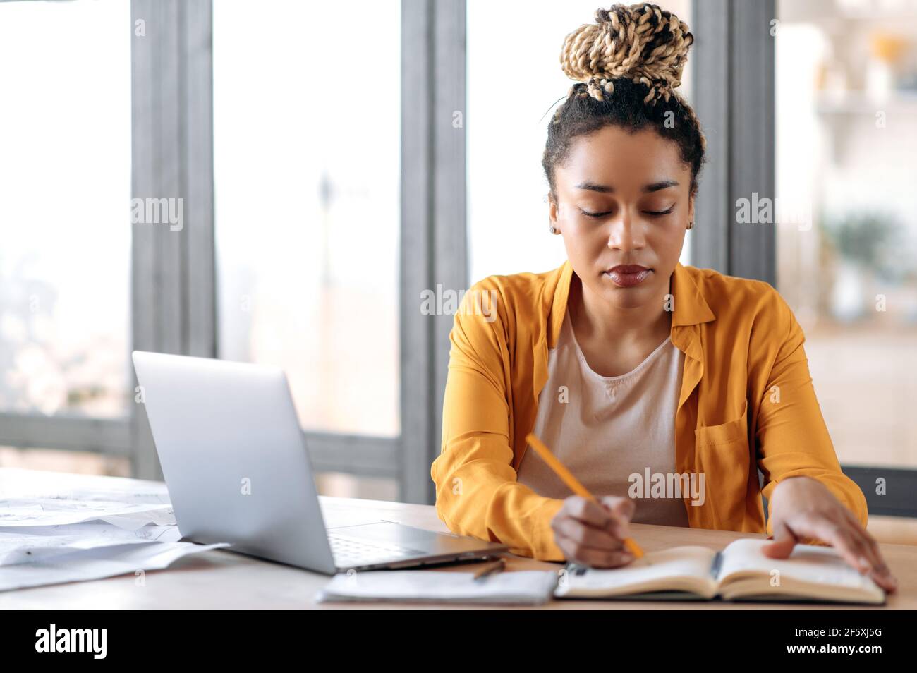 Focalisé élégant afro-américaine fille avec des dreadlocks, étudiante, dans des vêtements décontractés, étudier à la maison utilise un ordinateur portable, regarder des cours en ligne, prendre des notes dans un ordinateur portable, s'assoit dans le salon Banque D'Images